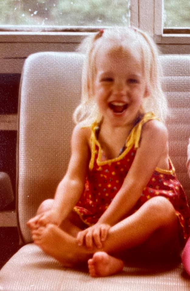 1970s girl with blond hair wearing a red sunsuit and laughing