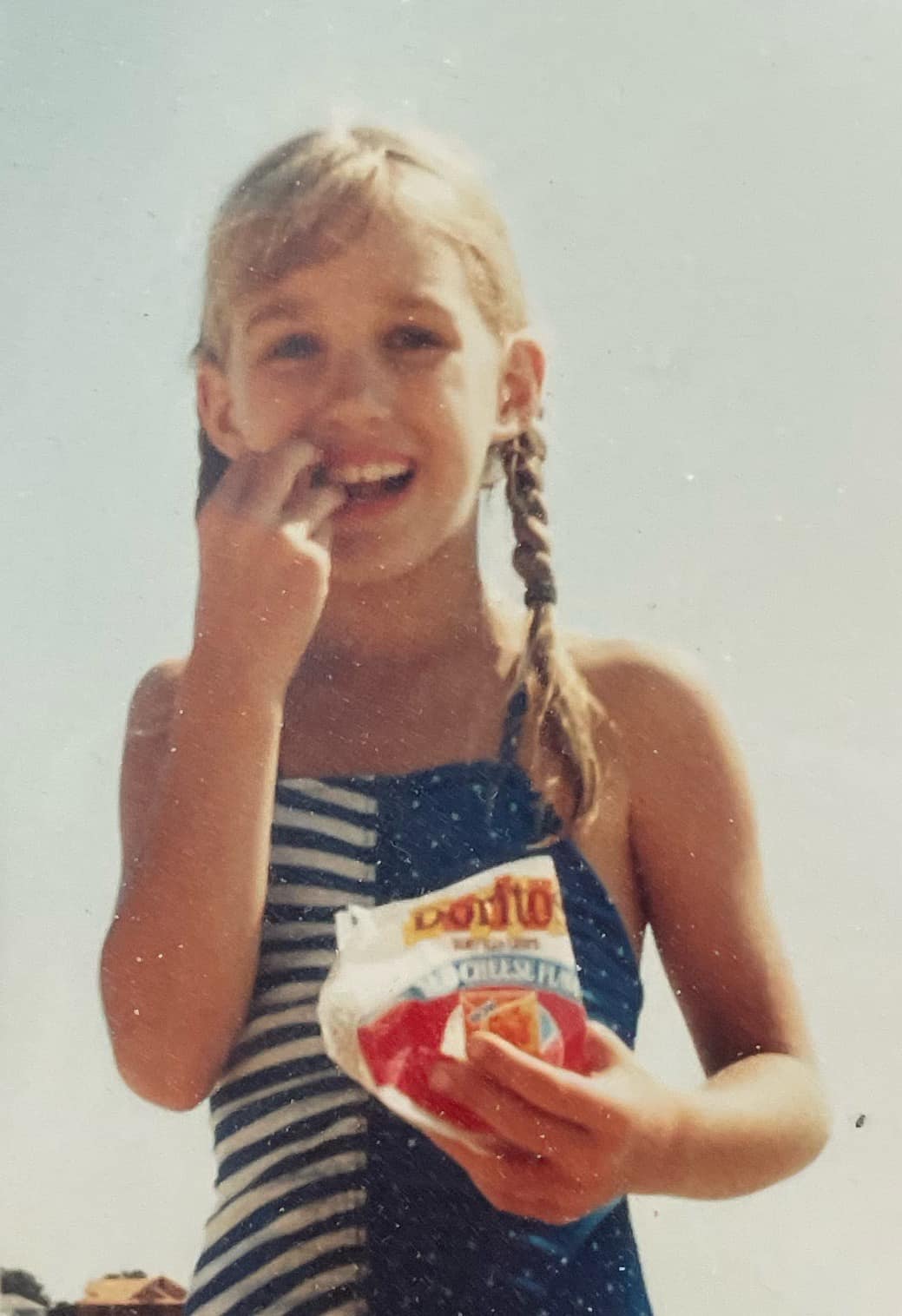 Young girl in 1984 eating a Dorito while smiling and holding the chip bag. She's wearing a blue and white one-piece bathing suit and wearing her blond hair in two braids.