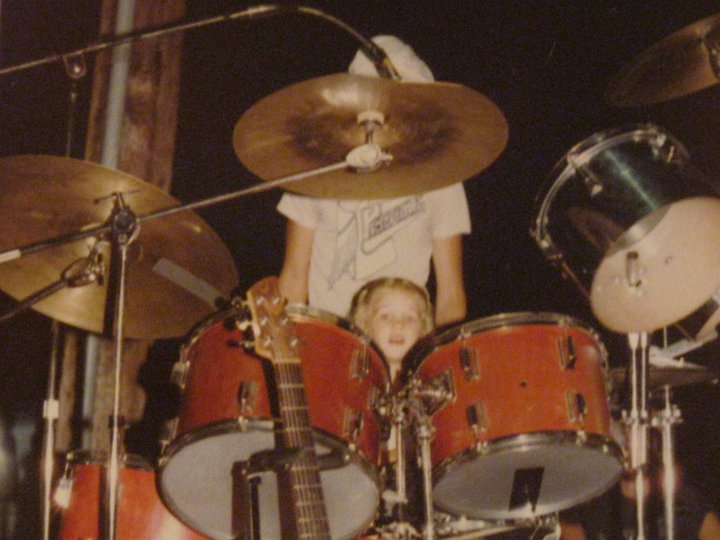 1970s little girls with blond hair sitting behind a drum set with only her head visible; her father is standing behind her, his face blocked by a cymbal.