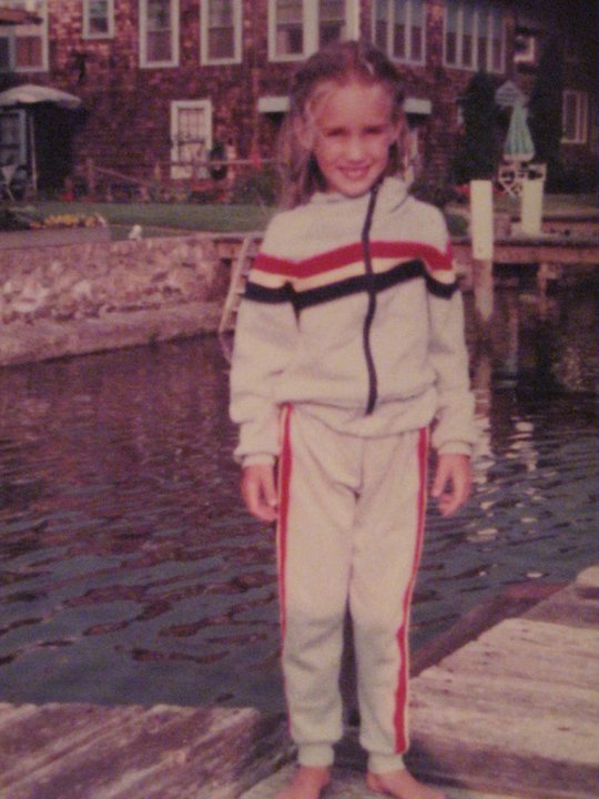 1980s girl with blond pigtails wearing a gray tracksuit that has blue, red, and yellow stipes across the chest and down each pant leg.