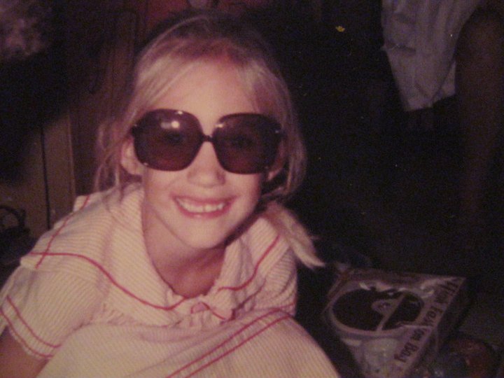 1980s girl with blond hair in a ponytail wearing large brown sunglasses and smiling