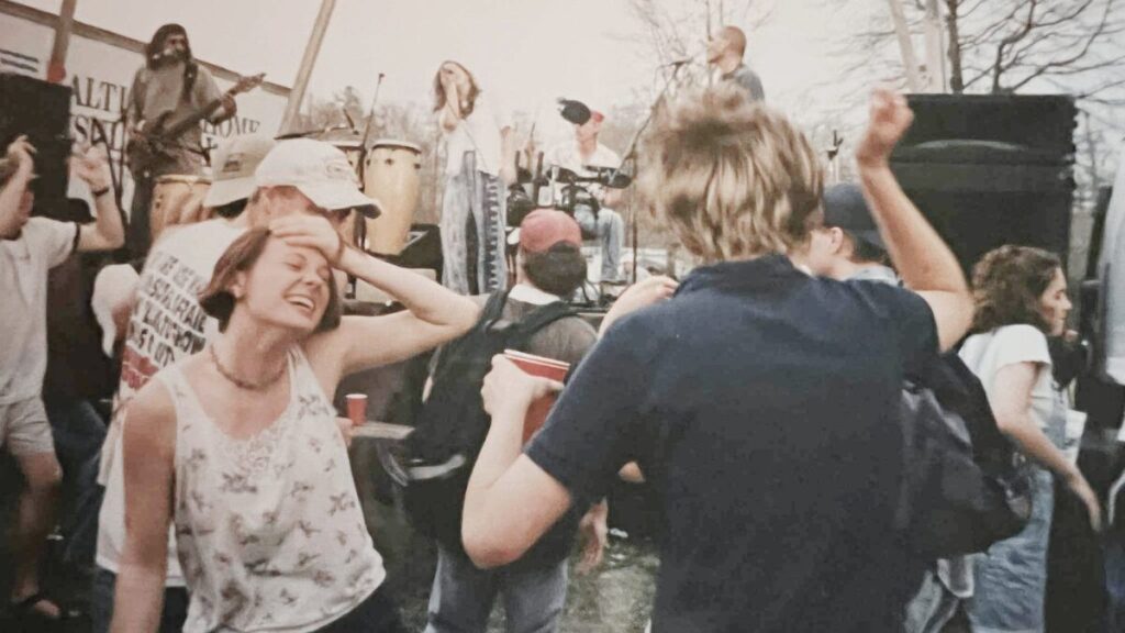 eople dancing near the stage at a 1990s outdoor music festival, with musicians performing in the background.