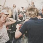 eople dancing near the stage at a 1990s outdoor music festival, with musicians performing in the background.