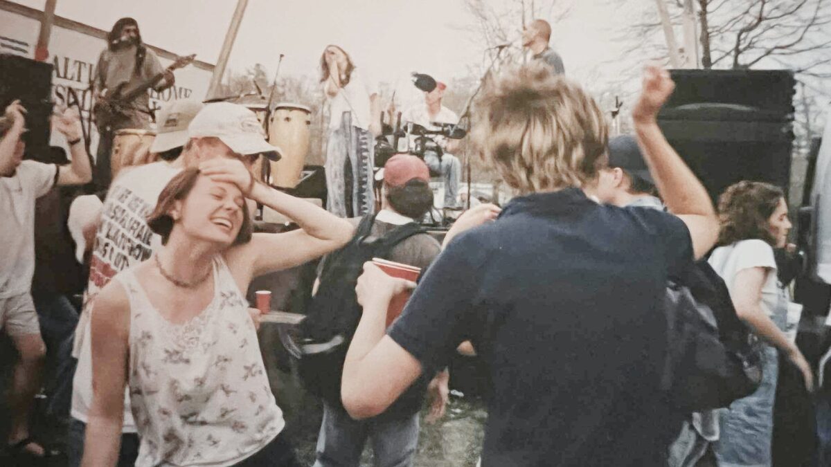 eople dancing near the stage at a 1990s outdoor music festival, with musicians performing in the background.