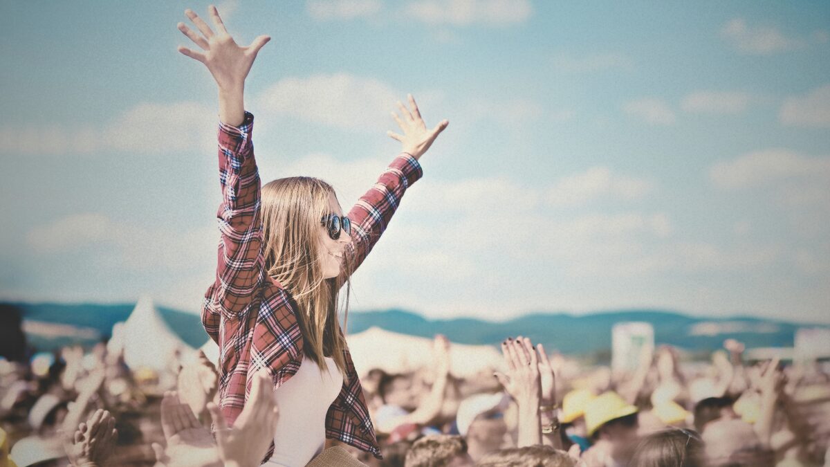 Crowd at a 1990s-style outdoor music festival with a woman in flannel on someone’s shoulders, hands raised in celebration.