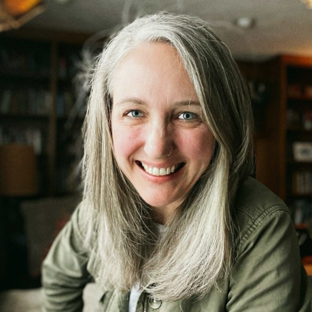 Close-up portrait of a smiling woman with long silver hair and wearing a green jacket, looking at the camera.
